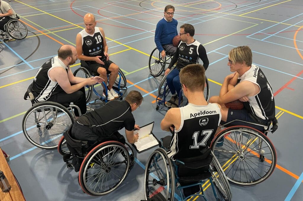 Meerdere rolstoel basketballers in een sporthal die overleg hebben, met de trainer die in zijn papieren kijkt